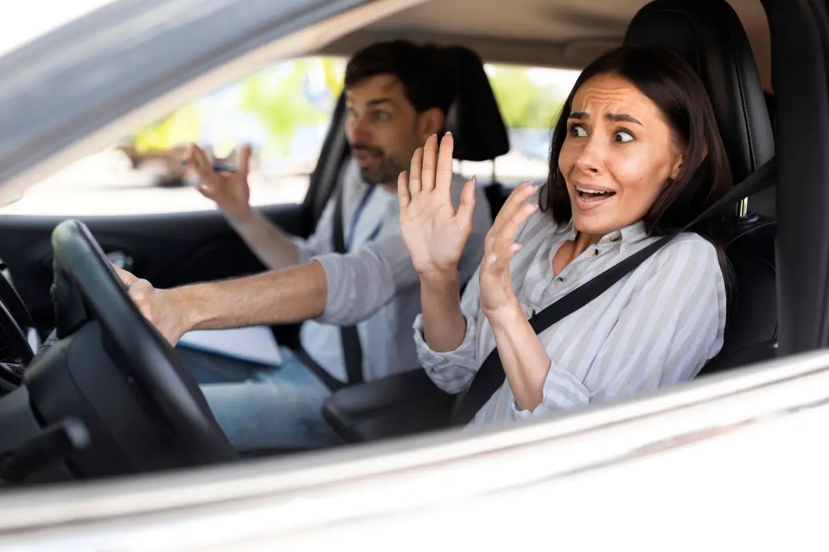 Woman driver is mock frightened by an mock irate male passenger who grabs the wheel.